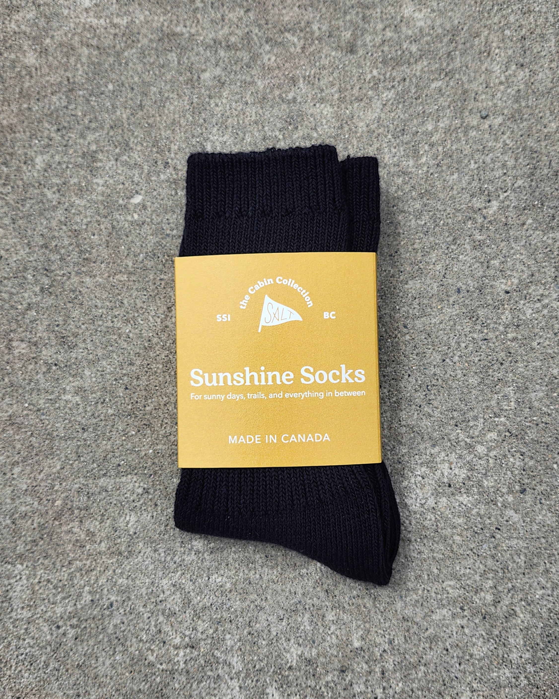 A pair of black ribbed crew socks with a tan paper label wrapped around them, placed on a textured gray surface. The label reads 'Sunshine Socks' and 'MADE IN CANADA'.
